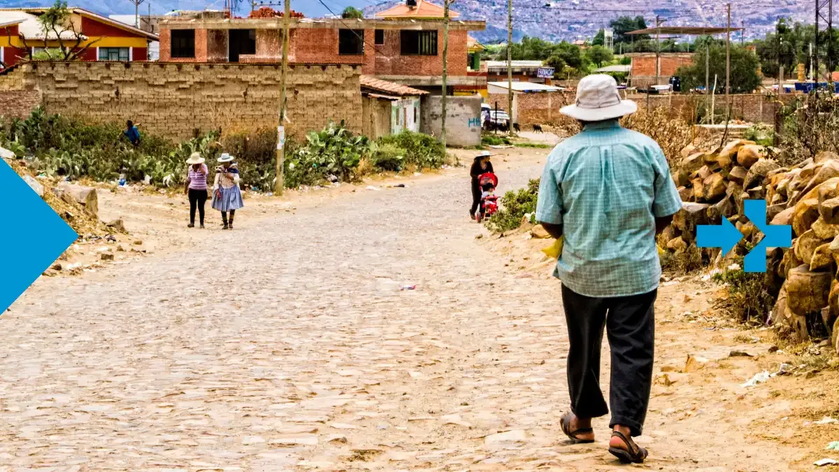 Hombre caminando por un camino empedrado en un pueblo rural con familias al fondo.