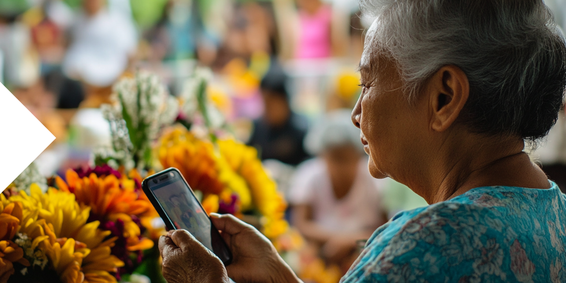 An elderly woman holding a phone with flowers in the background, remembering loved ones during a memorial event